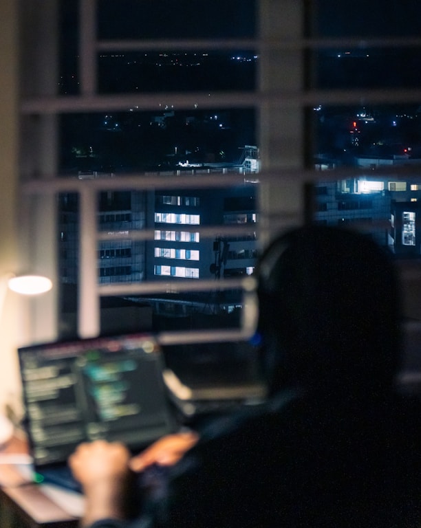 Person working on laptop at night with city view.