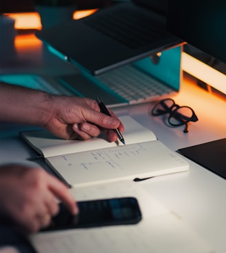 Hands writing in notebook near laptop and phone.