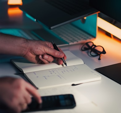 Hands writing in notebook near laptop and phone.